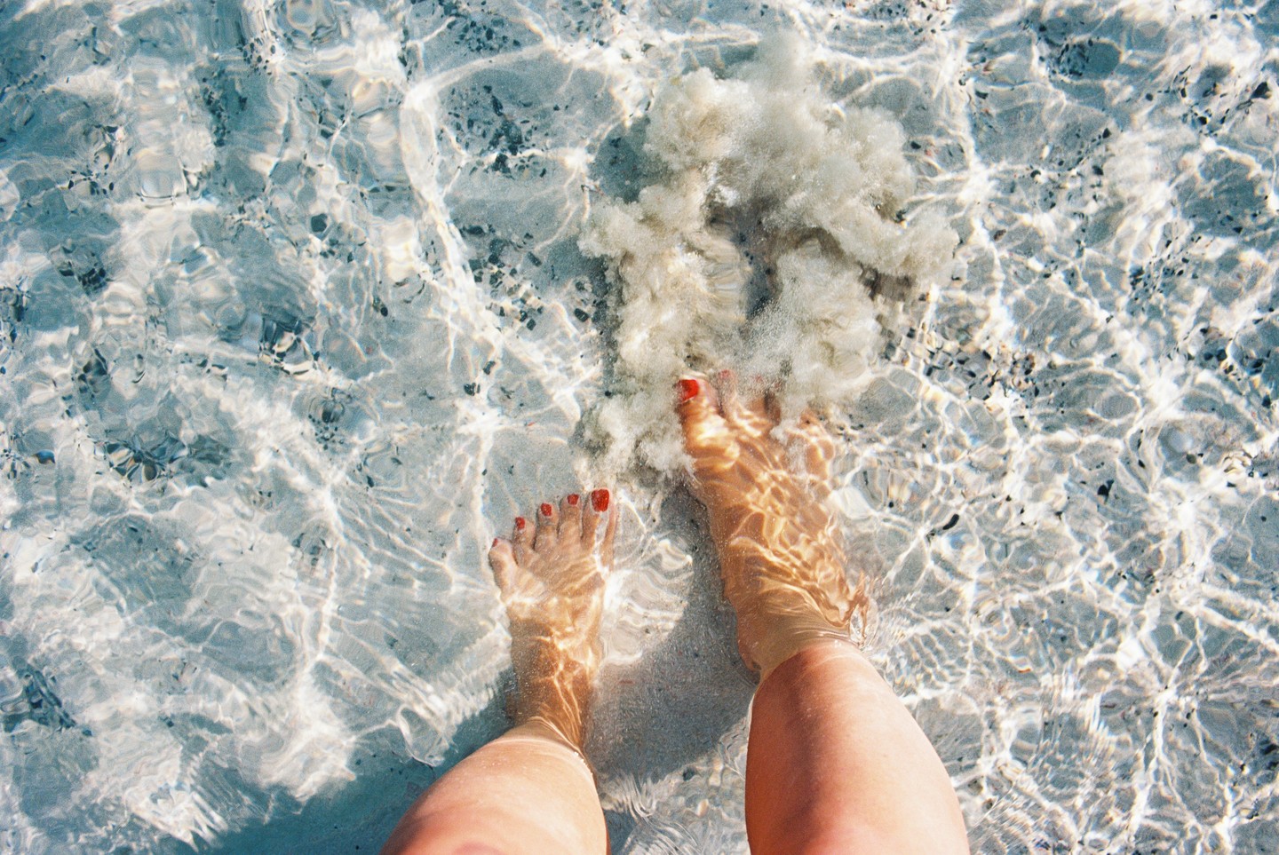 A person's legs and feet with red painted toenails are visible in clear shallow water with sand and small rocks on the bottom.
