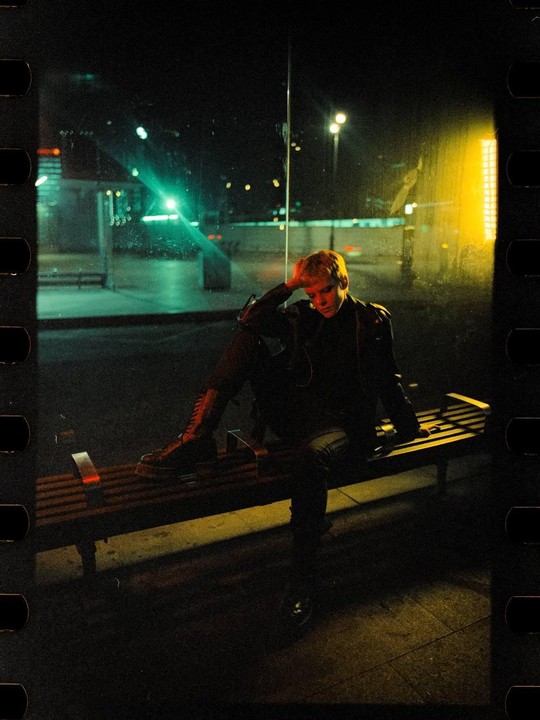 A person with short hair sits on a bench at a bus stop at night.