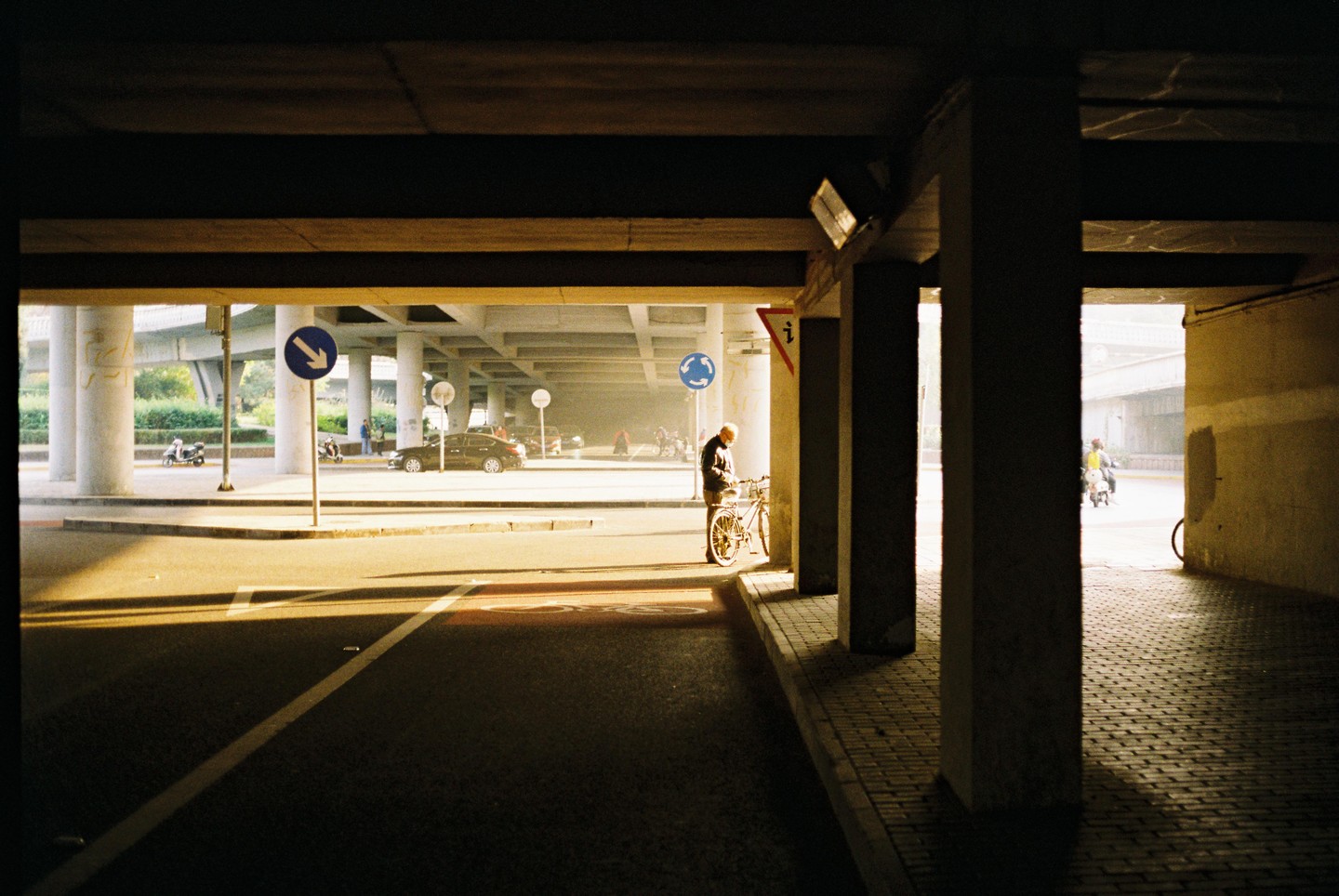 A man stands with his bicycle under a concrete overpass with cars and scooters in the background.