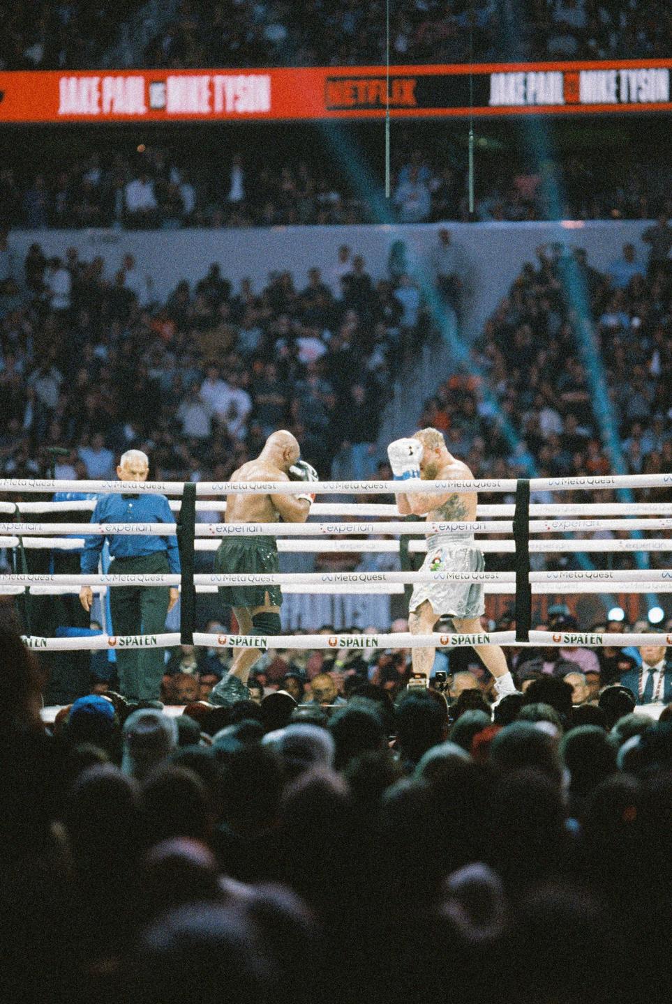 Two boxers and a referee are in a boxing ring surrounded by a large crowd.
