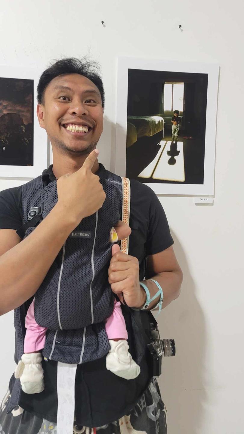 A man smiles at the camera while carrying a baby in a carrier and pointing to a framed photograph on a white wall.