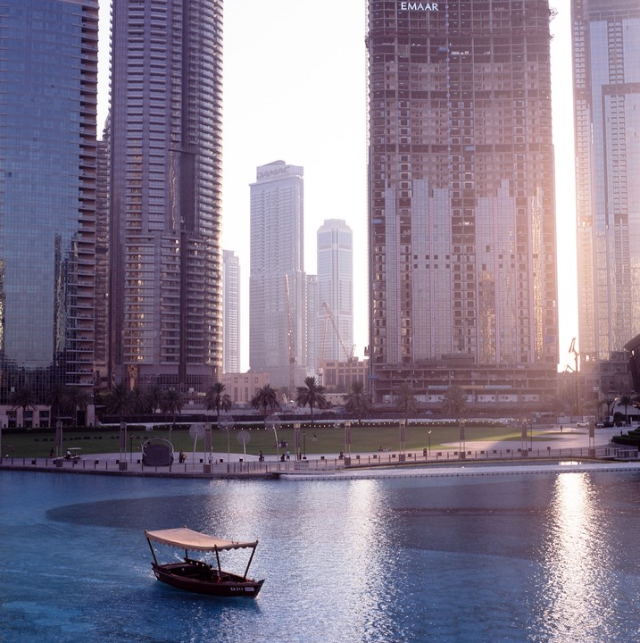 A small boat floats on blue water in front of a city skyline with tall buildings and palm trees.