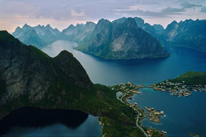 A small village with houses and boats is nestled between mountains and a body of water under a cloudy sky.