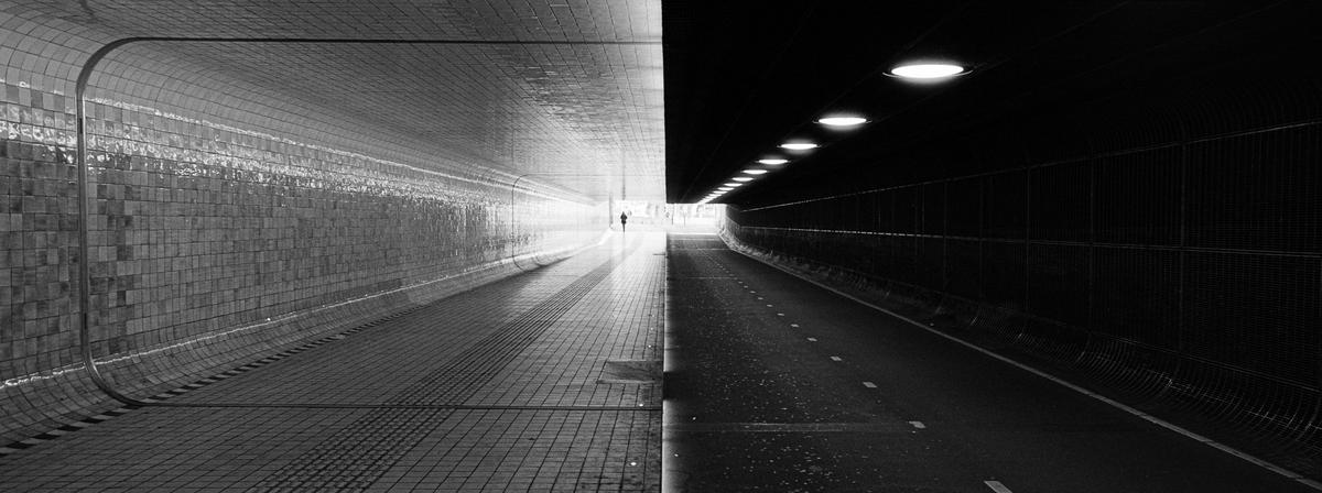 A person walks through a tunnel with a tiled wall on one side and a dark road on the other.