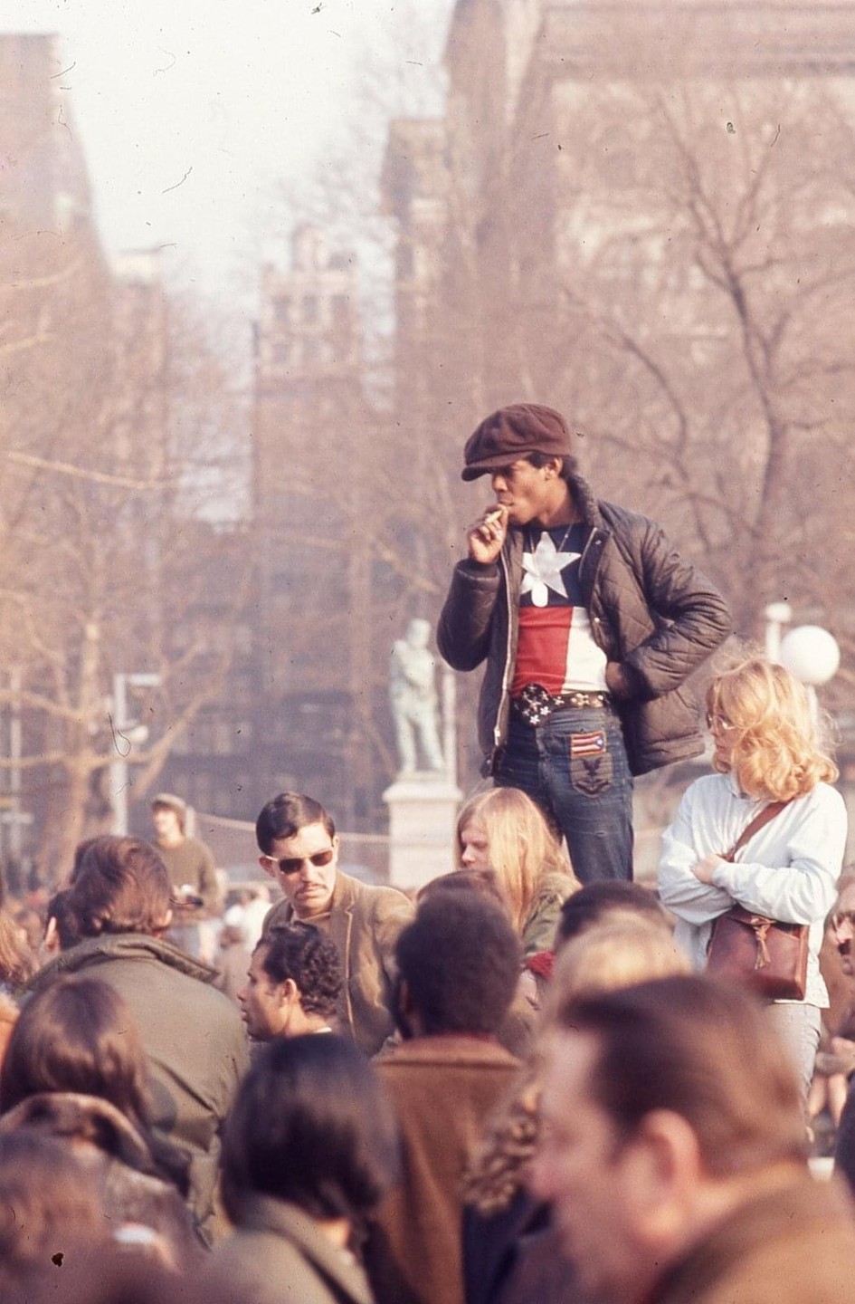 A man in a cap and a star-spangled shirt smokes a cigarette while standing above a crowd of people in Washington Square.