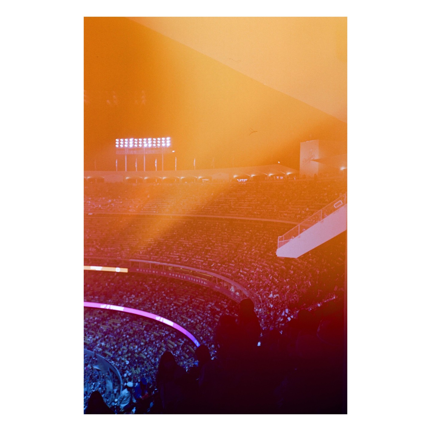 A light leak creates an orange glow over a stadium filled with spectators at night.