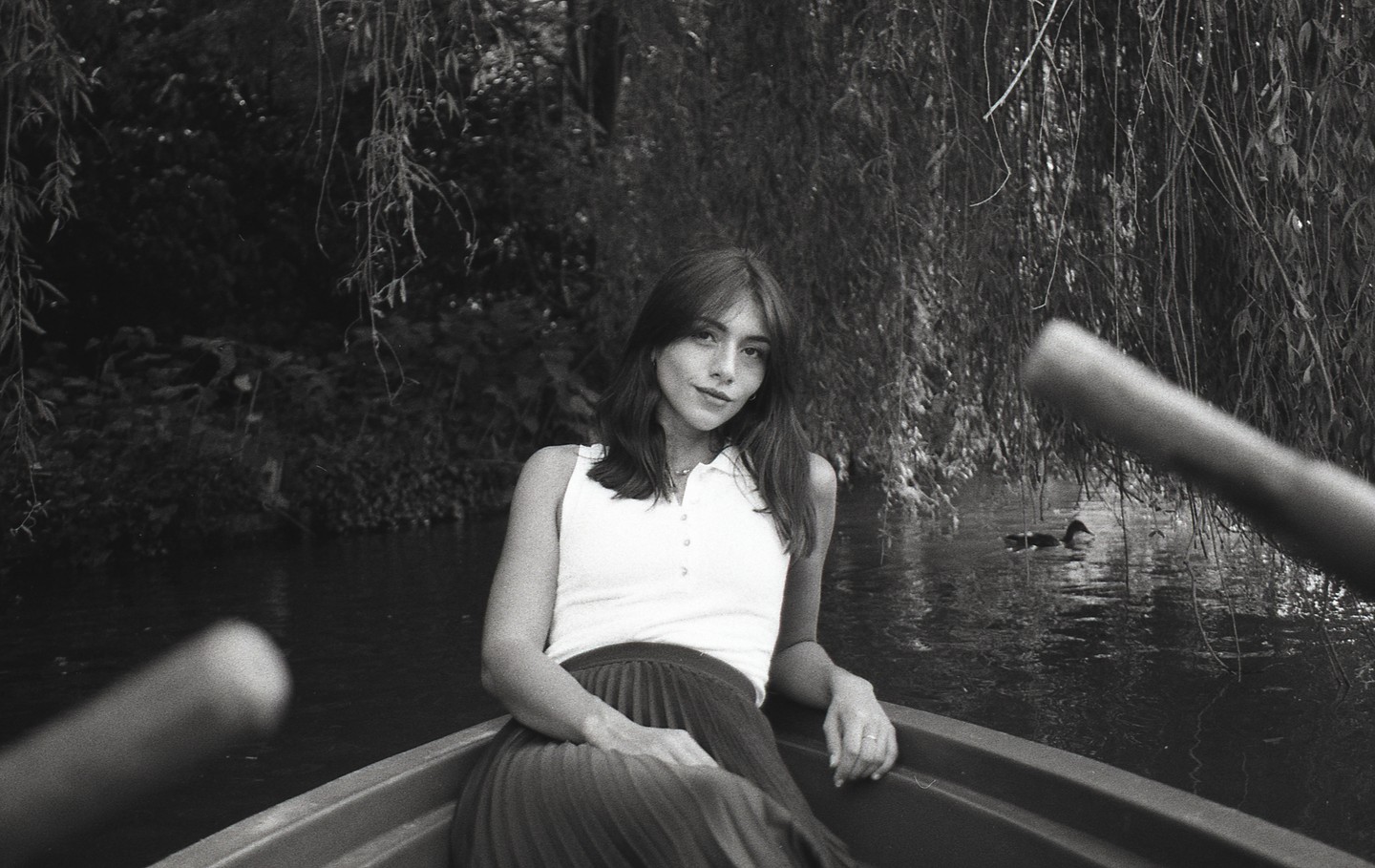 A woman sits in a rowboat on a river with weeping willow branches hanging over the water.