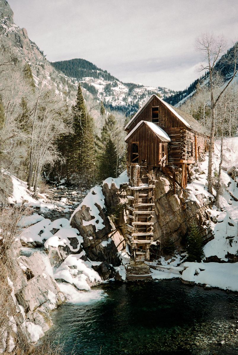 A wooden mill sits on a snowy cliff above a river with mountains in the background.