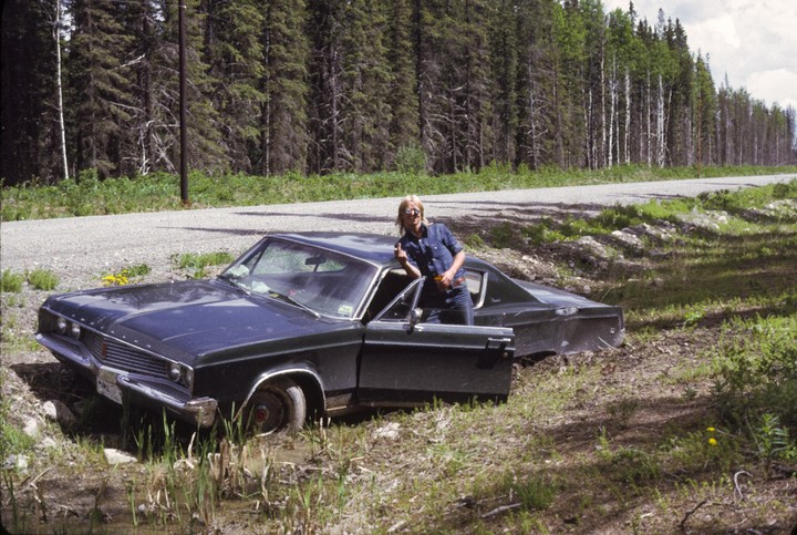 A man stands next to a dark car that is stuck in mud on the side of a dirt road, with a forest in the background.