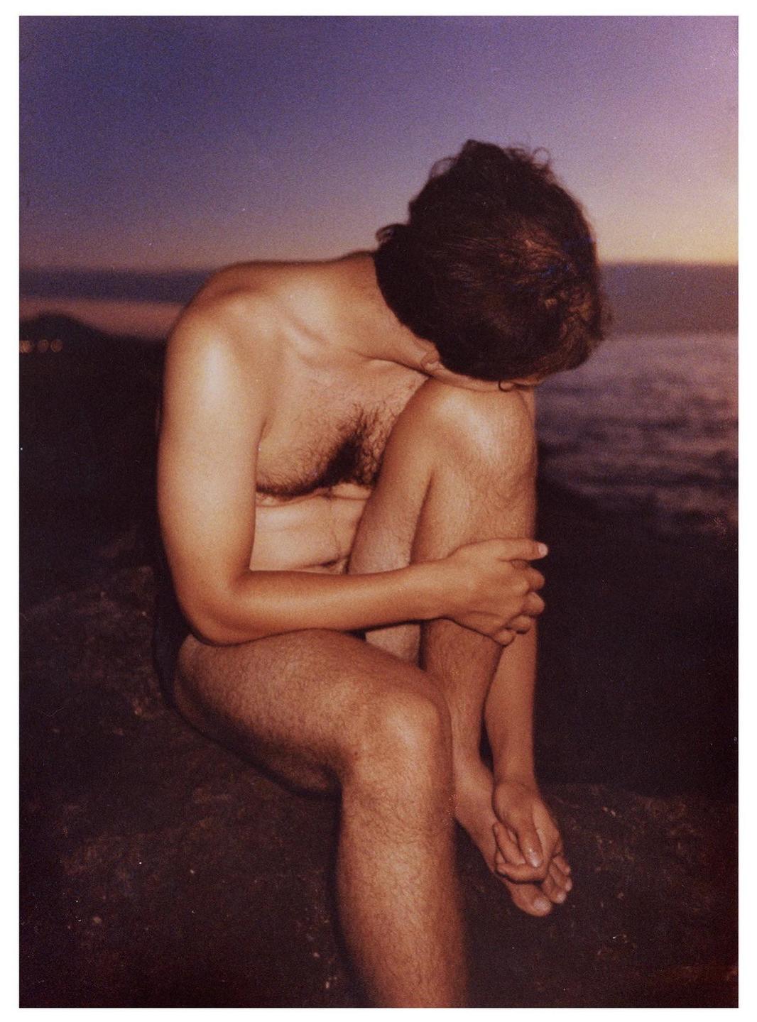 A man sits with his head bowed and arms wrapped around his knees by the water at dusk.