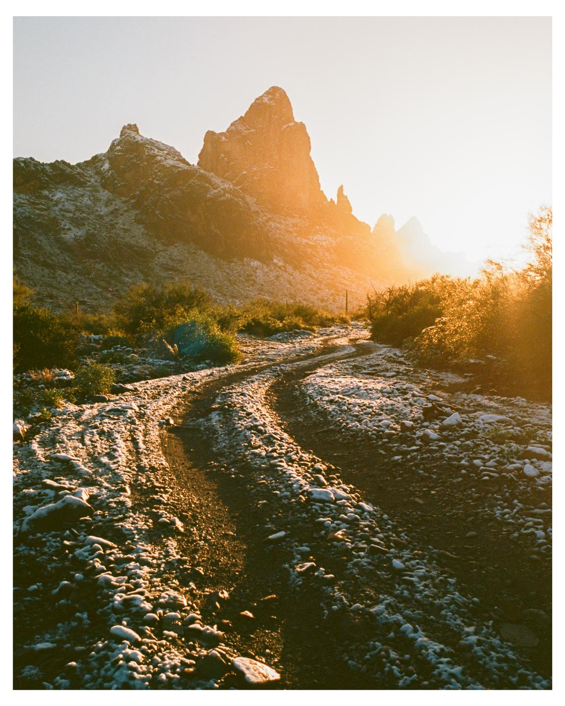 A dirt road covered in a dusting of snow leads towards a mountain range at sunset.