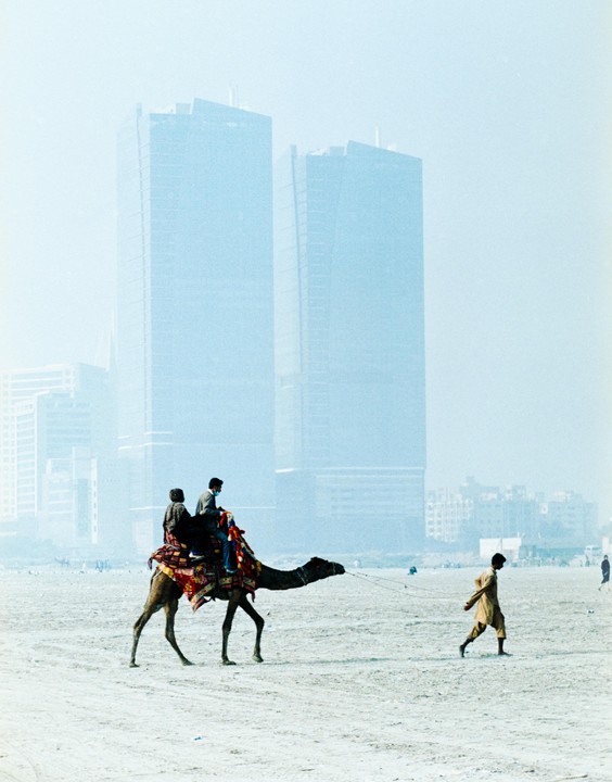 Two men ride a camel led by another man on a beach with tall buildings in the background.