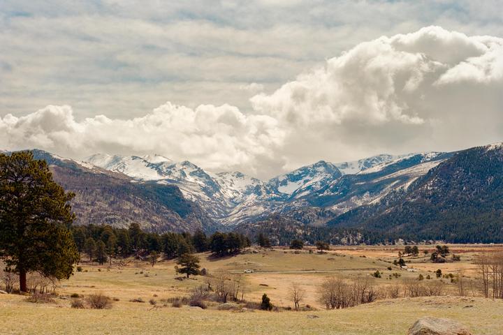 A vast landscape shows snow-capped mountains under a cloudy sky, with a valley of trees and fields in the foreground.
