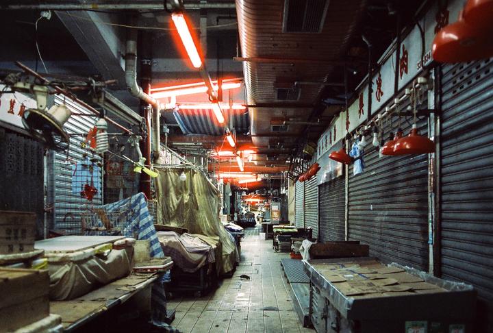A narrow market alley is illuminated by red neon lights and red lamps, with metal stalls and closed metal shutters lining both sides.
