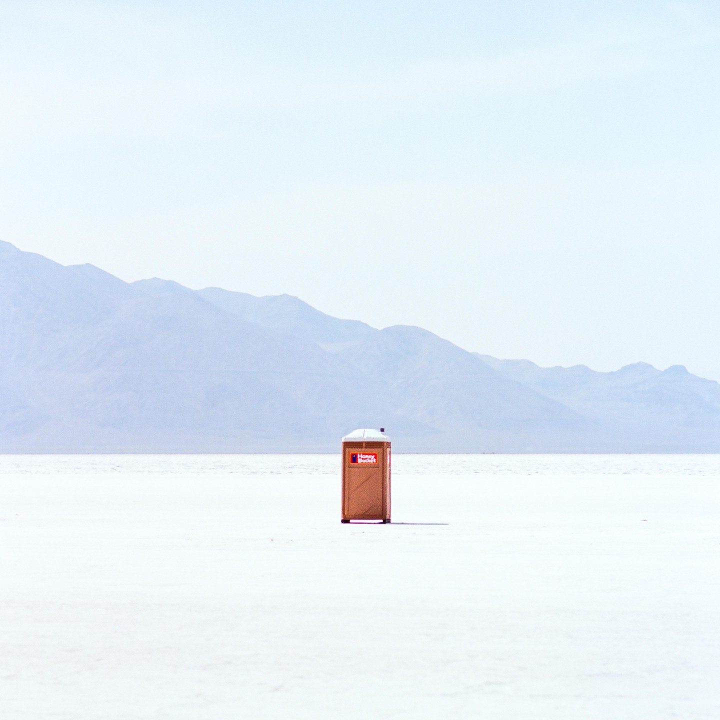 A portable toilet stands alone on a vast, flat, white expanse with mountains in the background.