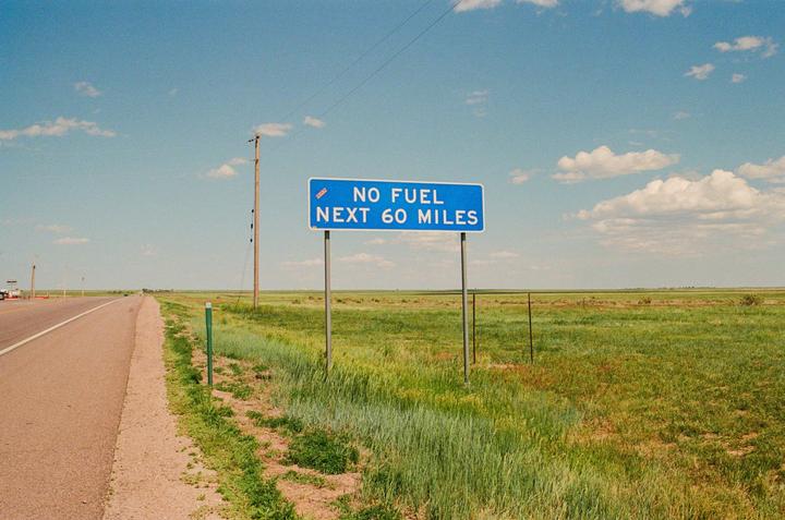 A blue road sign indicating "NO FUEL NEXT 60 MILES" stands beside a road with green fields under a blue sky with clouds.