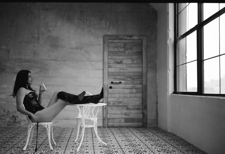 A woman in a corset and boots sits on a chair with her legs resting on a table, holding a drink in a room with a patterned floor and a large window.