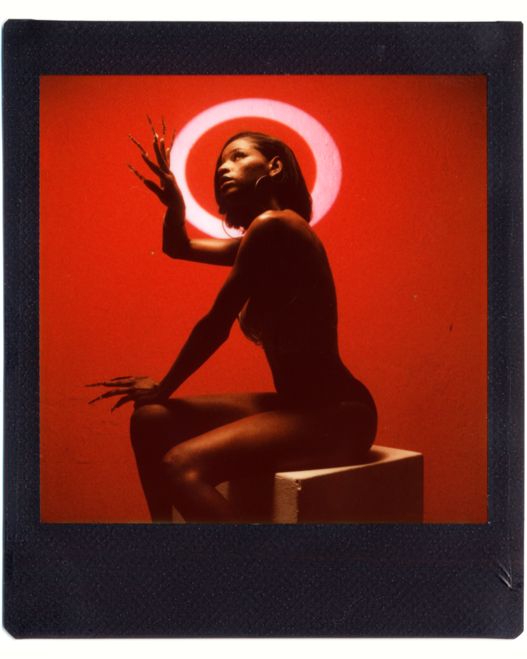 A woman with long nails and hoop earrings sits on a white block against a red background with a white circle.