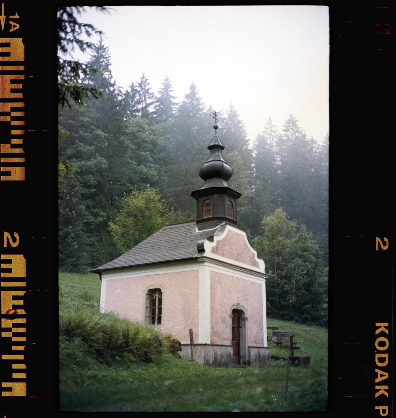 A small pink chapel with a dark dome and cross stands on a grassy hill with a forest in the background.