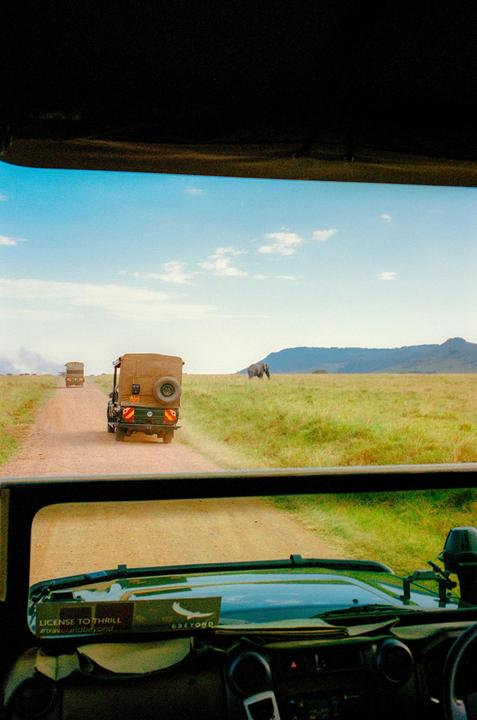 A safari vehicle drives on a dirt road with an elephant in the distance and another safari vehicle ahead.
