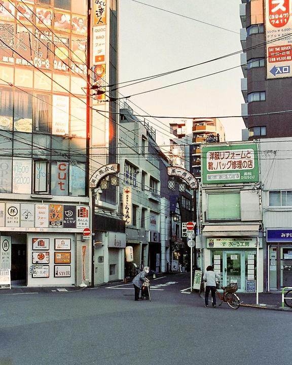 Two people are on a quiet street corner in a Tokyo neighborhood with buildings and power lines overhead.