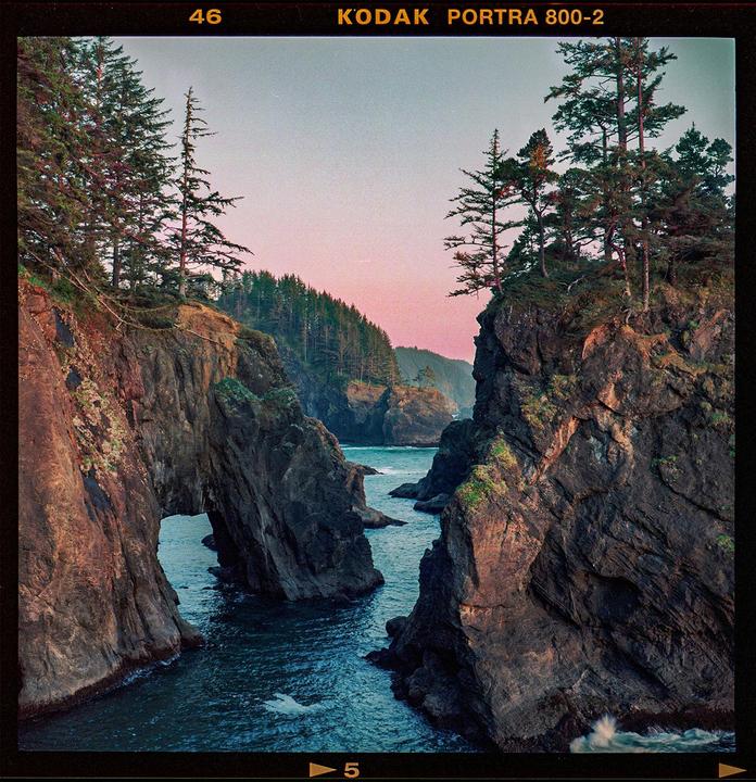 A body of water flows between two rocky cliffs with pine trees on top under a pink and blue sky.
