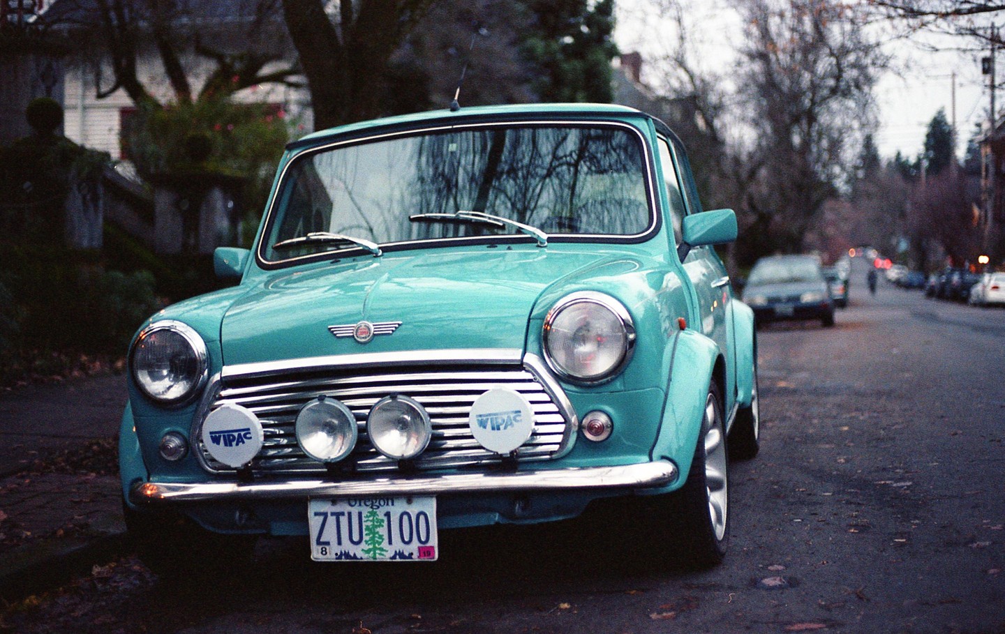 A turquoise Mini Cooper is parked on a street lined with trees and other cars.
