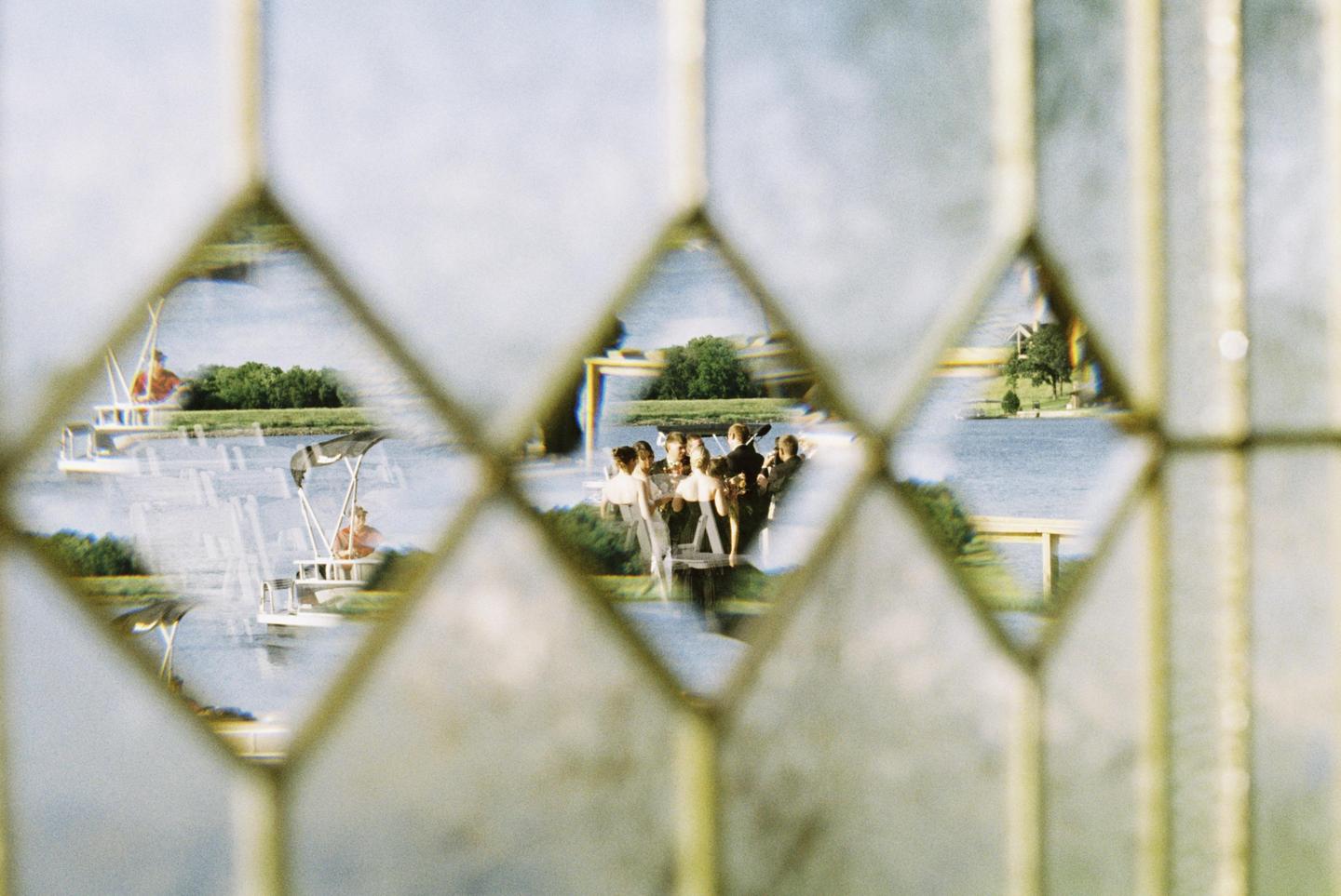 A group of people are visible through a diamond-patterned window overlooking a body of water with boats.