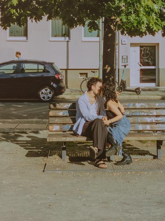 A couple sits on a park bench and kisses under a tree on a sunny day.