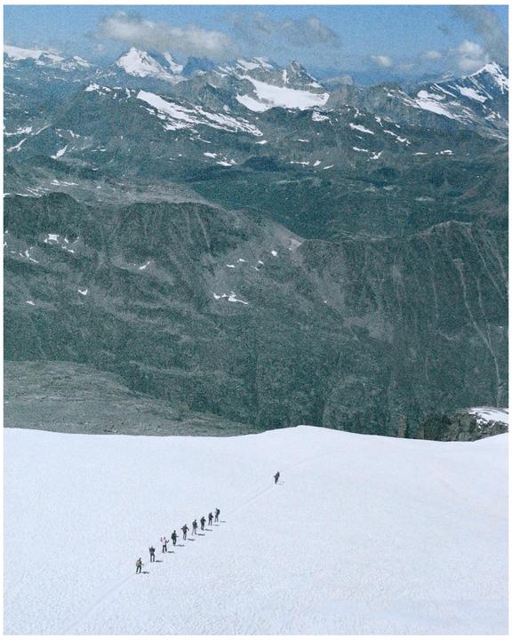 A group of people walk in a line across a snow-covered mountain with a vast mountain range in the background.