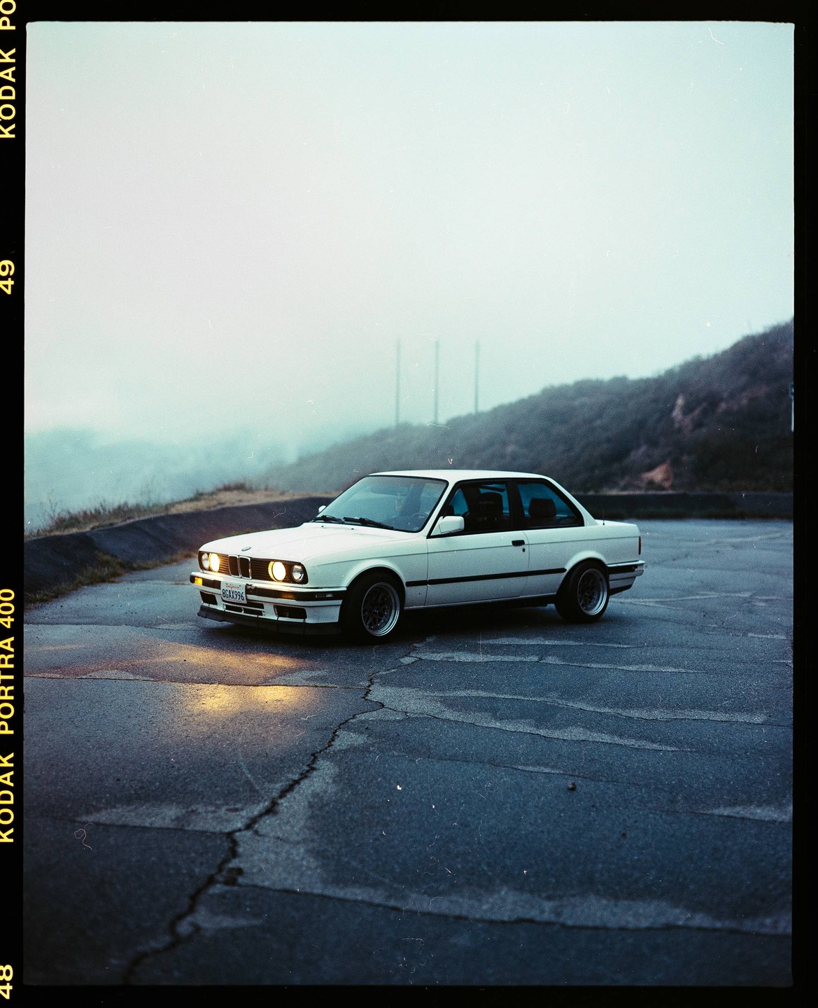 A white car with its headlights on is parked on a cracked asphalt surface with a hazy hill in the background.