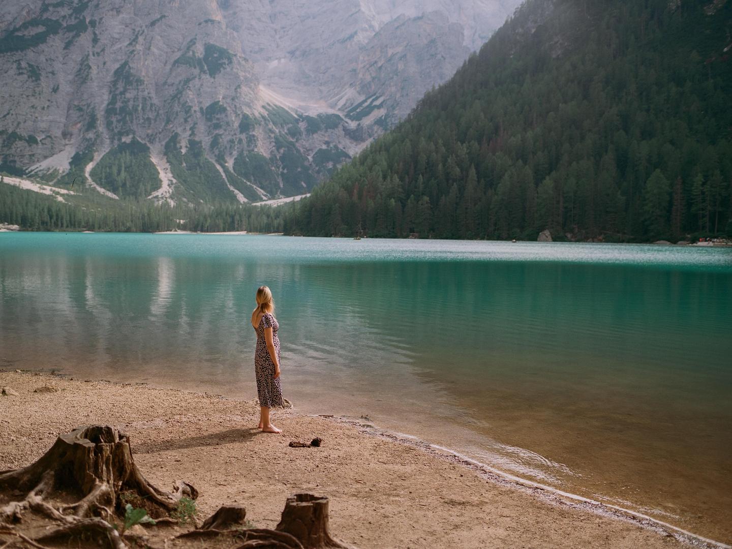 A woman stands barefoot on the shore of a turquoise lake, looking out at the water and mountains.