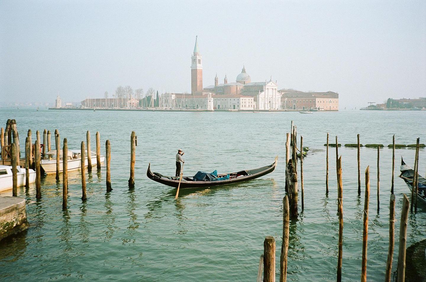 A gondolier stands in a gondola on the water with a large building and bell tower in the background.