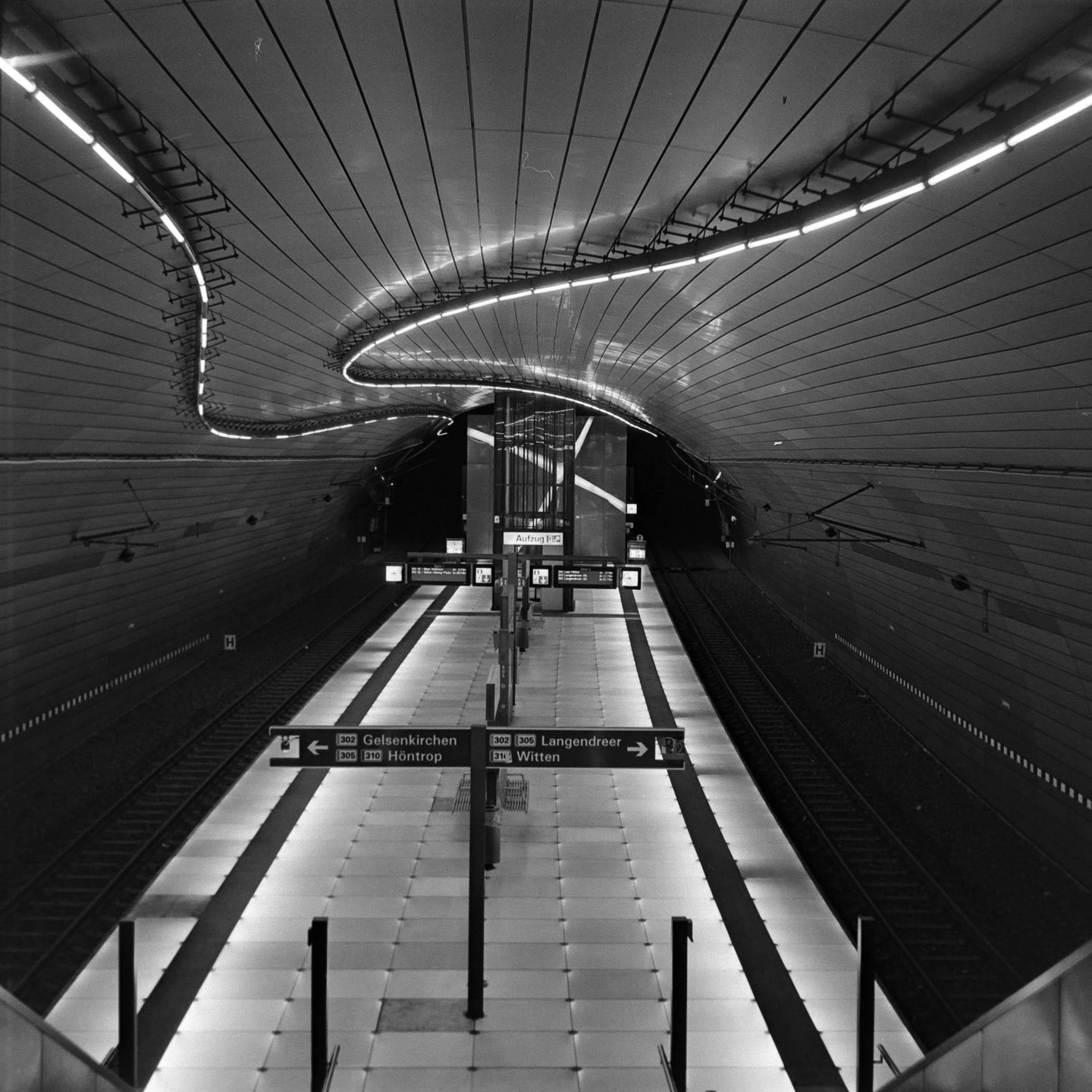 A black and white photograph shows a subway station with a curved ceiling and tracks on either side of a platform.