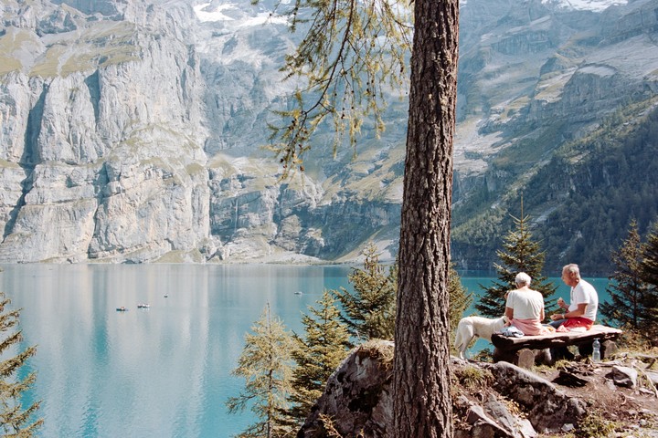 A couple and their dog sit on a wooden bench overlooking a lake surrounded by mountains.
