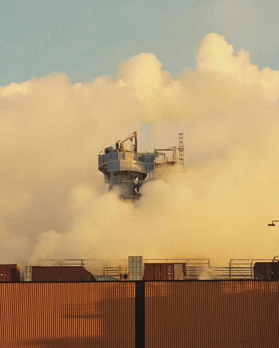 A factory building with a tall structure is partially obscured by a large cloud of smoke.