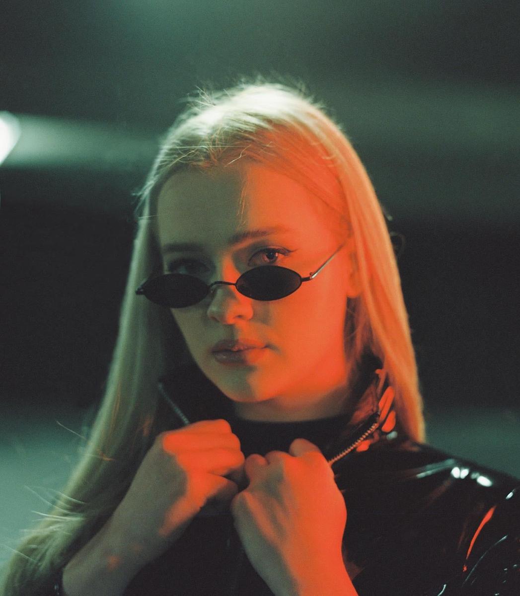 A woman with long blonde hair and sunglasses holds the collar of her jacket.