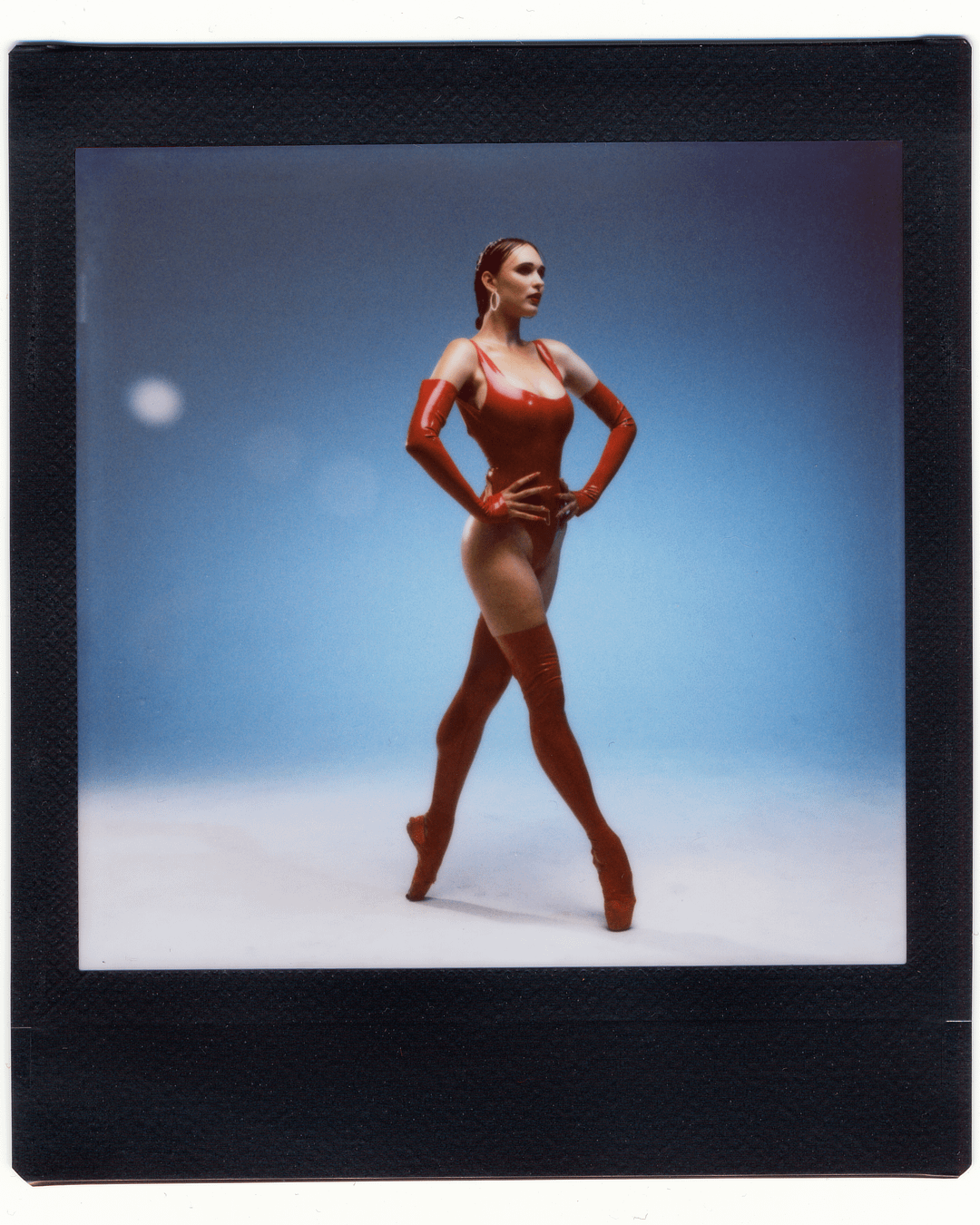 A woman in a red latex bodysuit and red pointe shoes poses with her hands on her hips against a blue background.