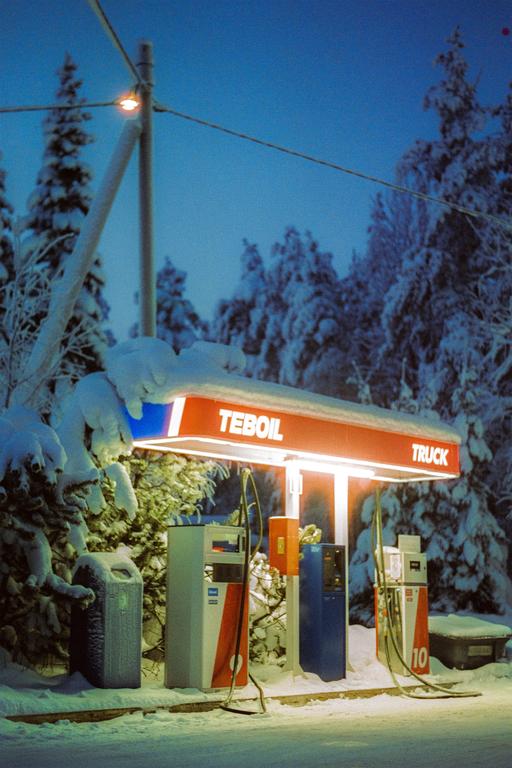 A Teboil gas station with a red and white canopy stands illuminated at night, surrounded by snow-covered trees and ground.