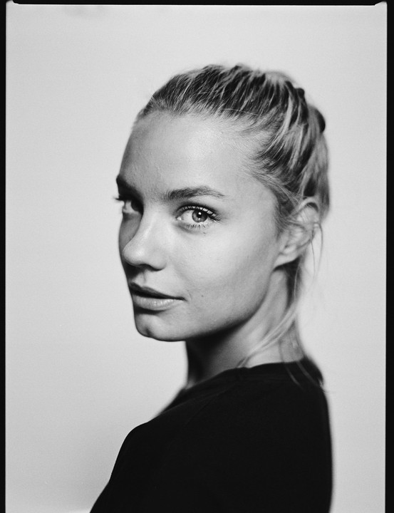 A woman with her hair pulled back looks over her shoulder at the viewer in a black and white portrait.