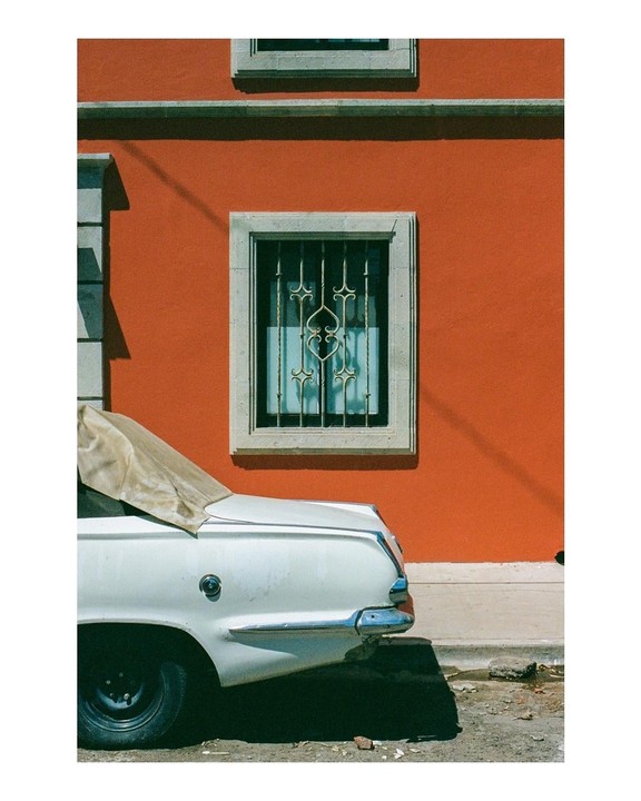 A white car with a tan convertible top is parked next to a vibrant orange building with a window featuring an ornate metal grate.