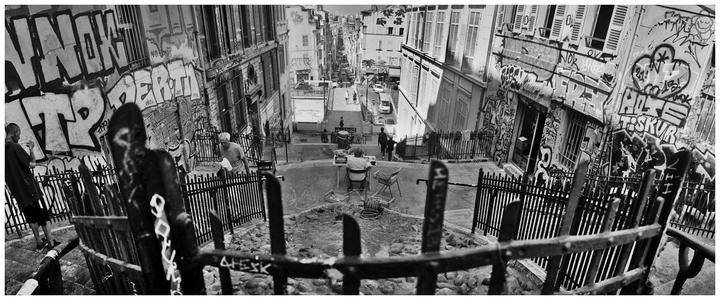 A panoramic black and white street scene shows a person sitting at a table on a cobblestone street, surrounded by buildings covered in graffiti.