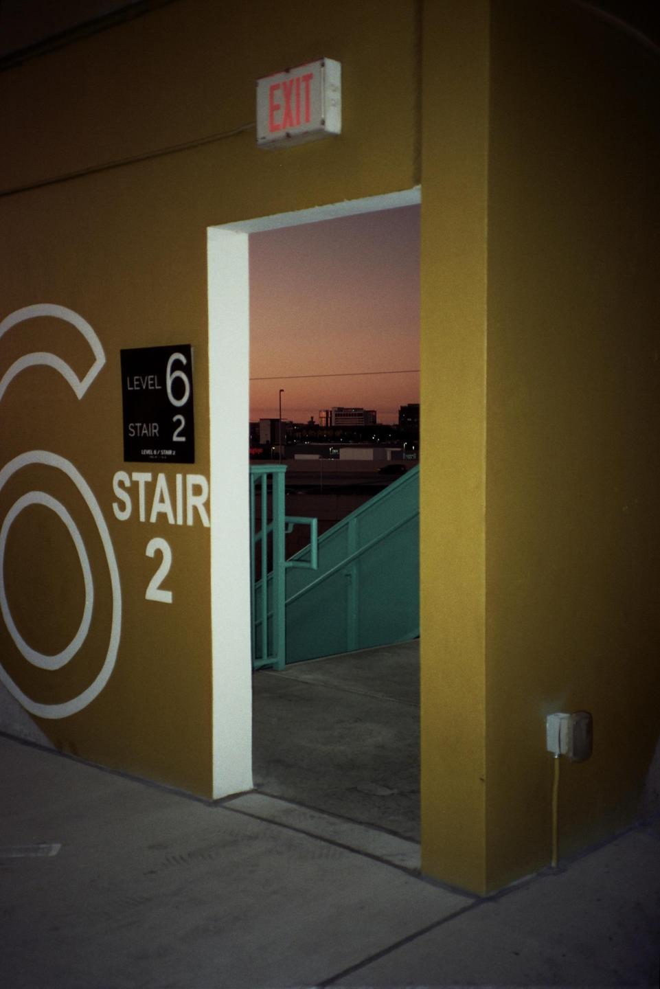 A yellow wall with a large white number six and a sign for level six, stair two, opens to a view of a teal staircase and a city skyline at dusk.