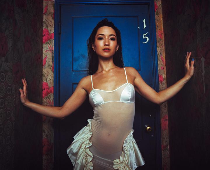 A woman in a sheer white outfit stands in front of a blue door with the number 15 on it, next to a wall with floral wallpaper.