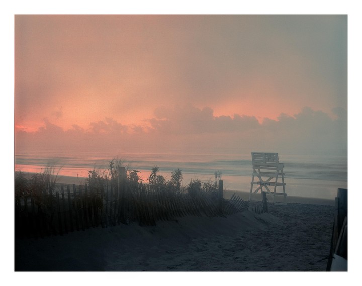 A lifeguard chair stands on a beach at sunrise, with a fence and dune grass in the foreground.