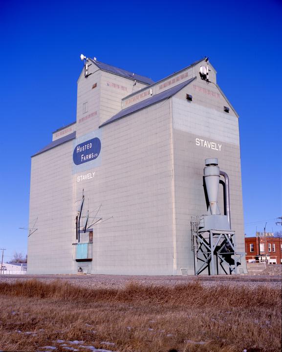 A large grain elevator with "Husted Farms Ltd" and "Stavely" written on its side stands under a clear blue sky.