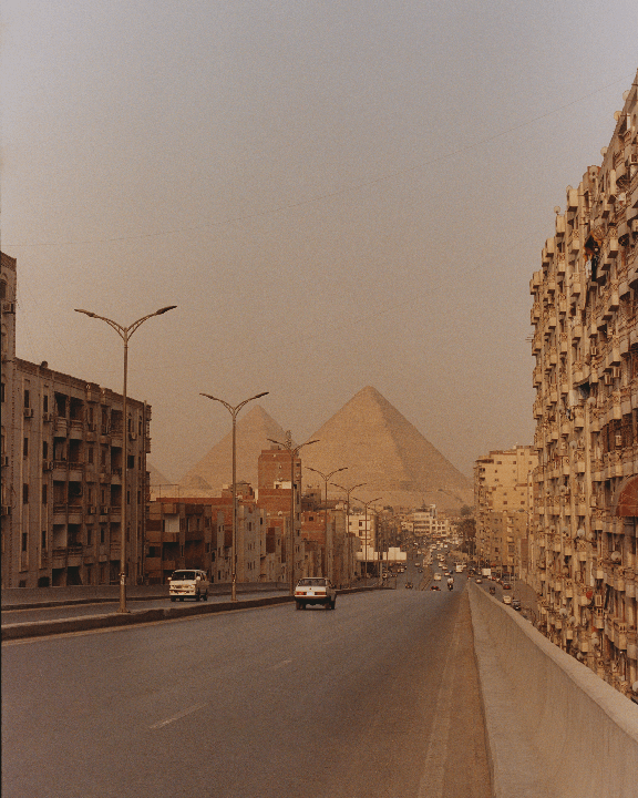 A wide road with cars and buildings on either side leads towards the pyramids in the distance.