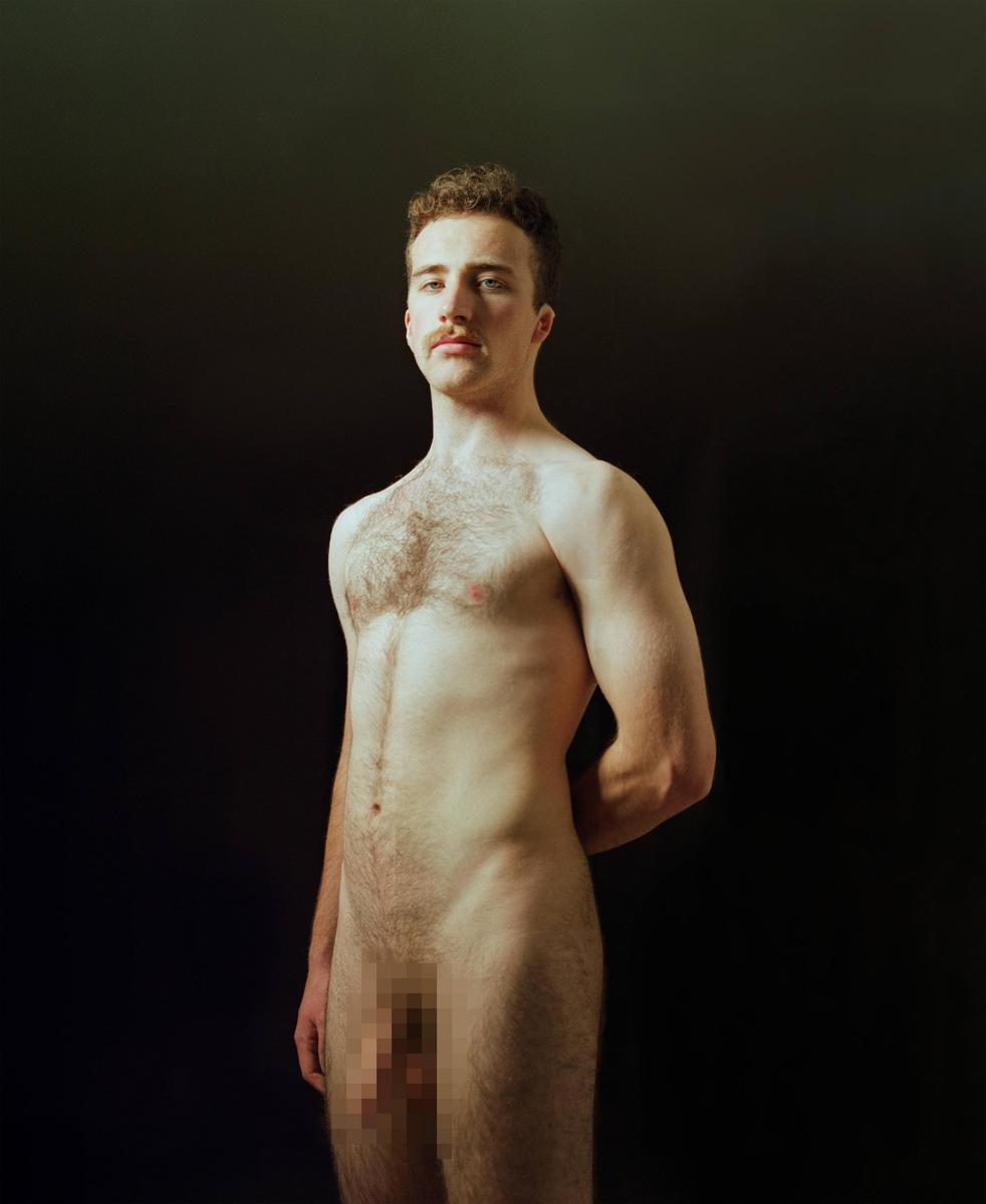 A man with a mustache stands nude against a dark background, looking directly at the viewer.