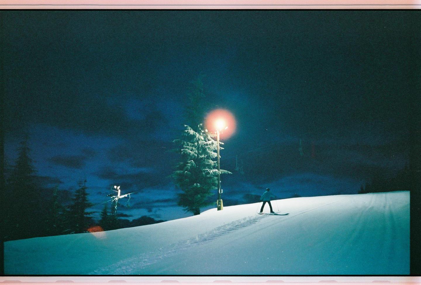 A skier ascends a snowy slope at night, illuminated by a bright light next to a pine tree.