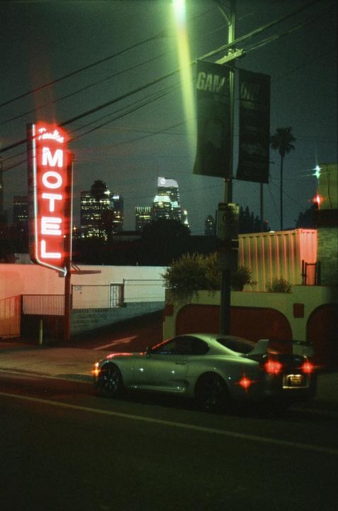 A silver sports car is parked on a street at night, with a neon motel sign and city lights in the background.